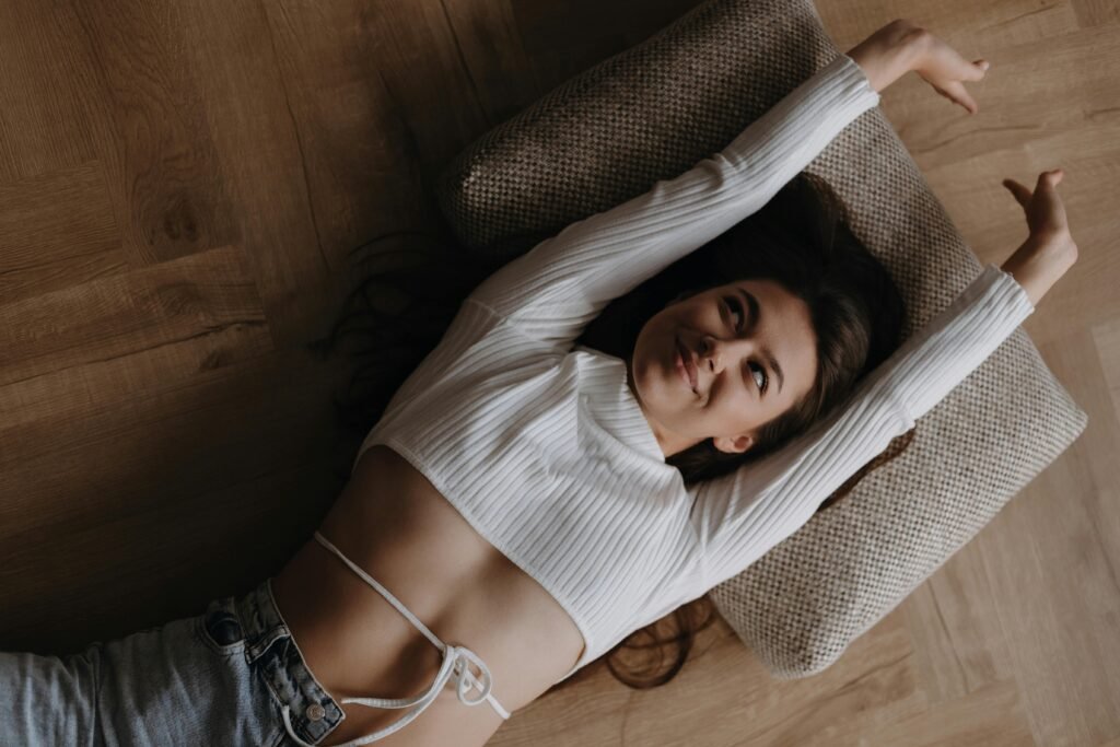 Young woman in a white crop top smiling while stretching on parquet floor with a cushion.